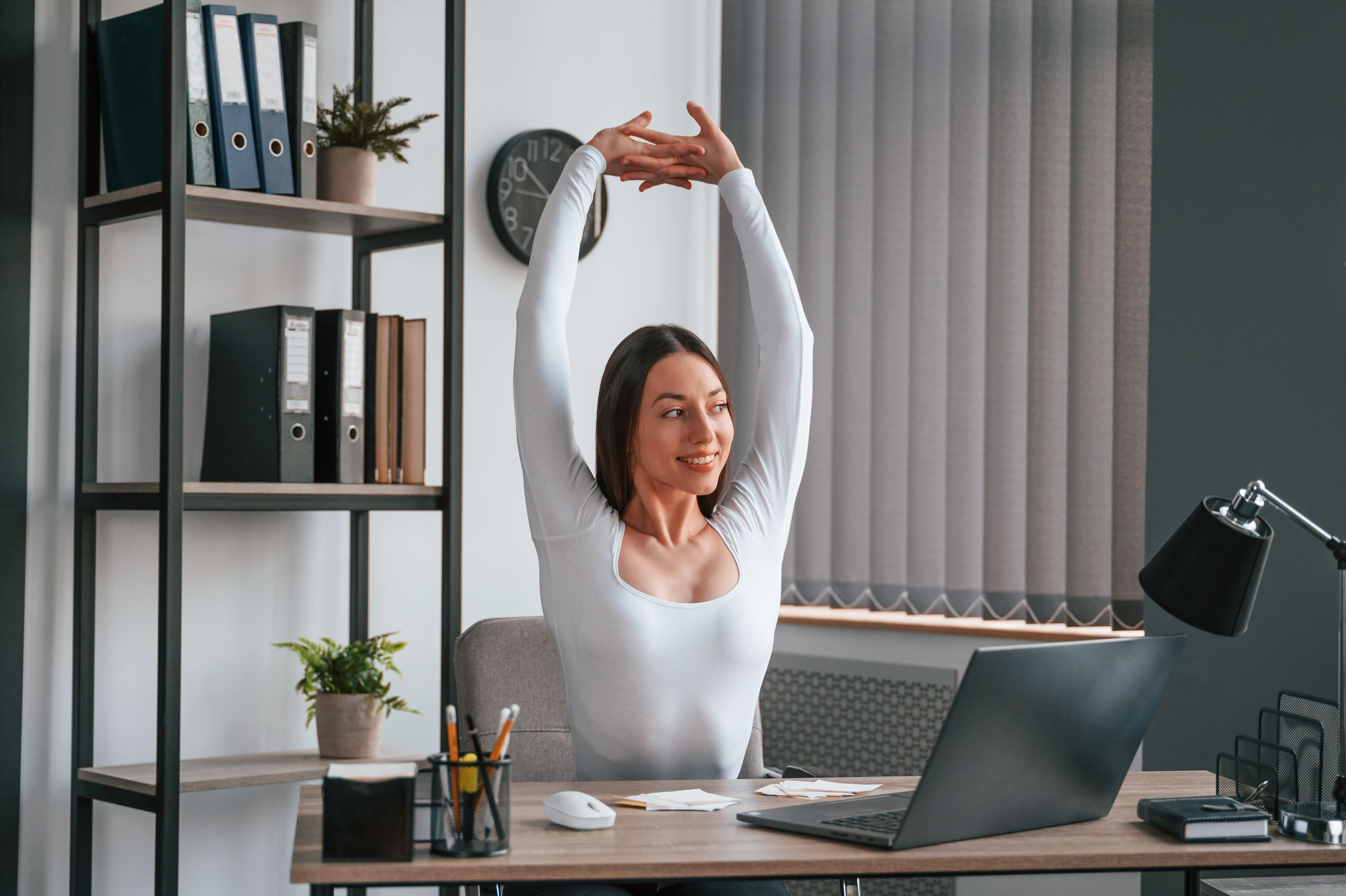 A professional woman stretching her arms behind her desk at work, taking a break to improve posture and relieve tension.